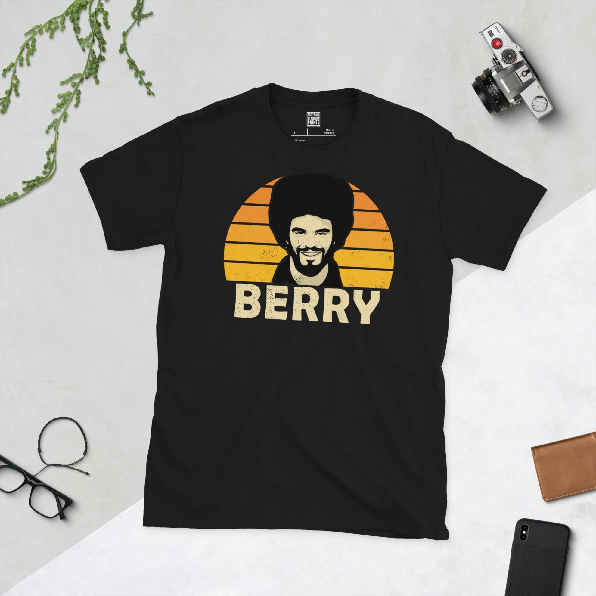 A black T-shirt featuring a stylised graphic of Wolverhampton Wanderers legend George Berry's profile, set against a Golden Orange striped sunburst background. The name "BERRY" is printed in light yellow below the portrait.