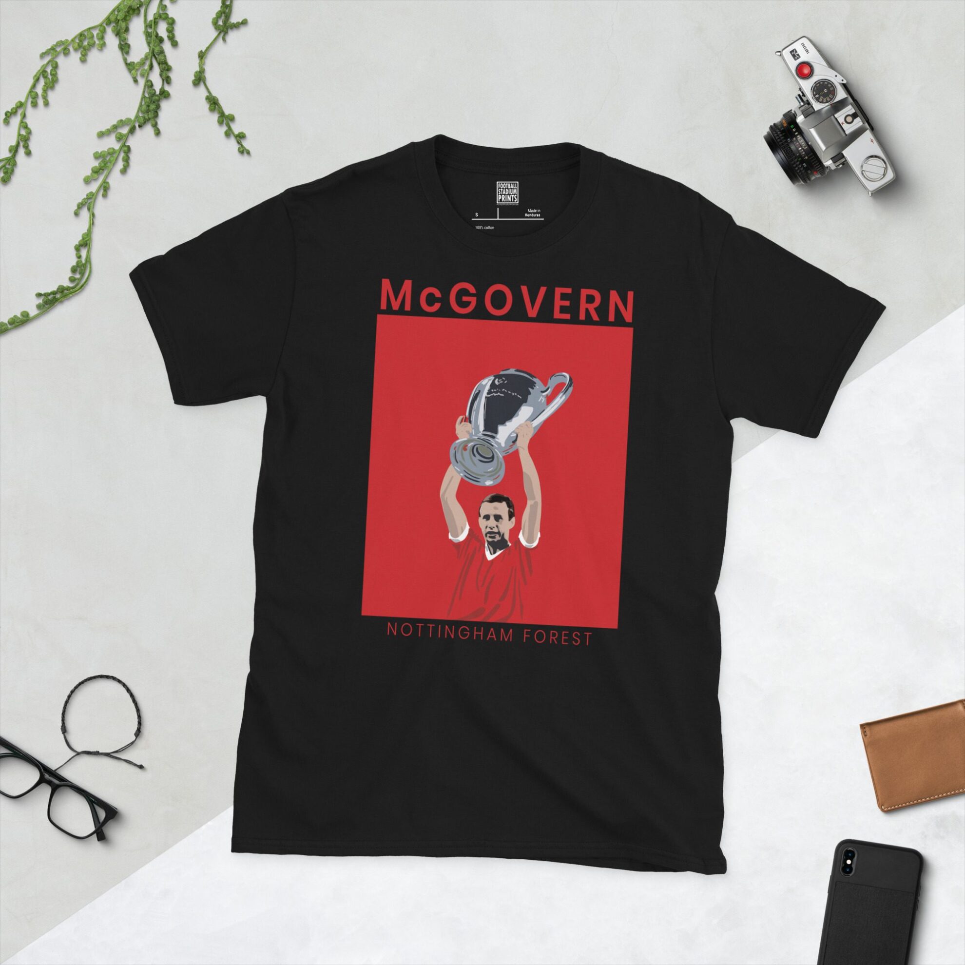 A black T-shirt featuring a solid white graphic of Nottingham Forest legend John McGovern lifting the European Cup trophy, set against a red and white background. The name "MCGOVERN" is printed in white below the portrait.