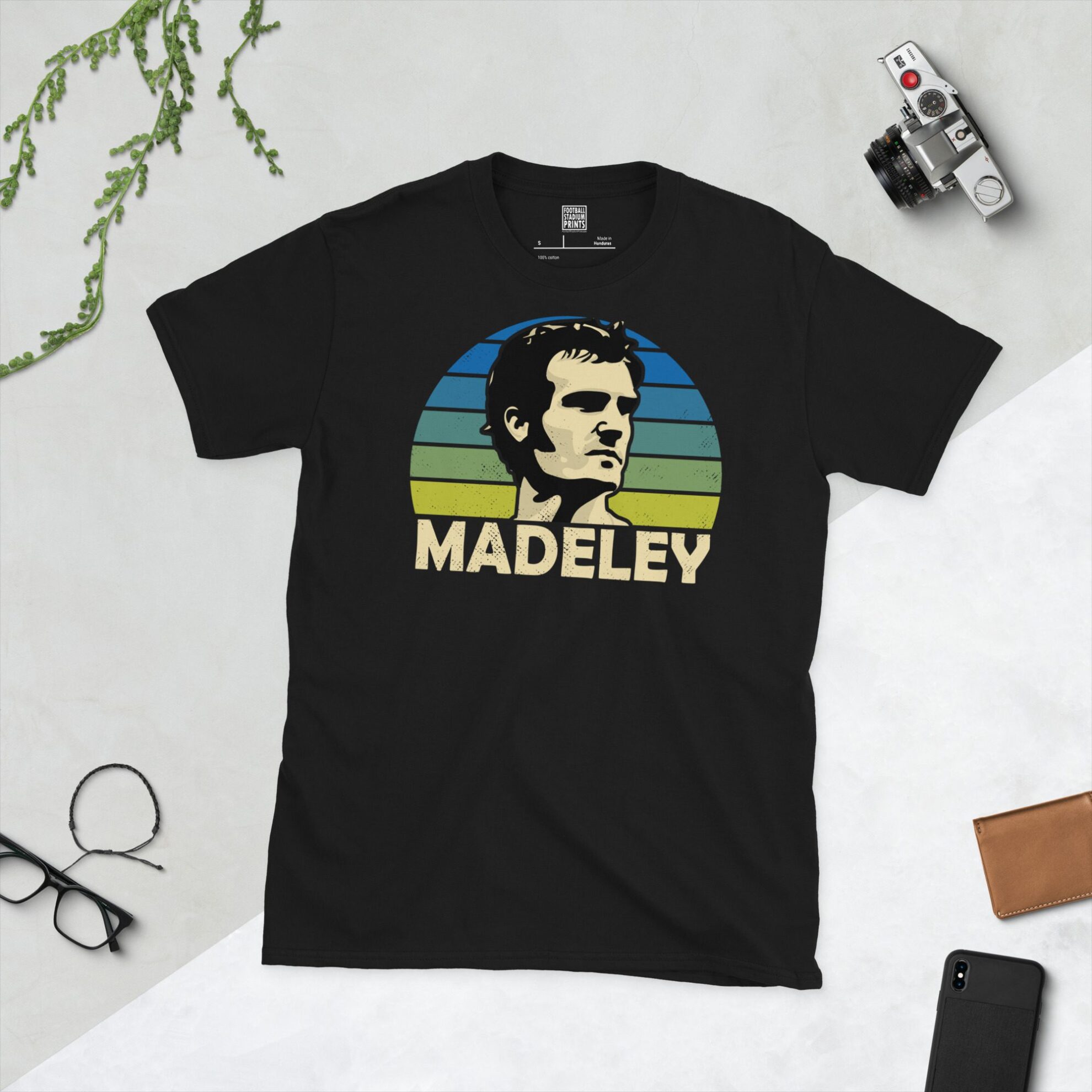 A black T-shirt featuring a stylised graphic of Leeds United legend Paul Madeley's profile, set against a blue to yellow faded striped sunburst background. The name "MADELEY" is printed in light yellow below the portrait.
