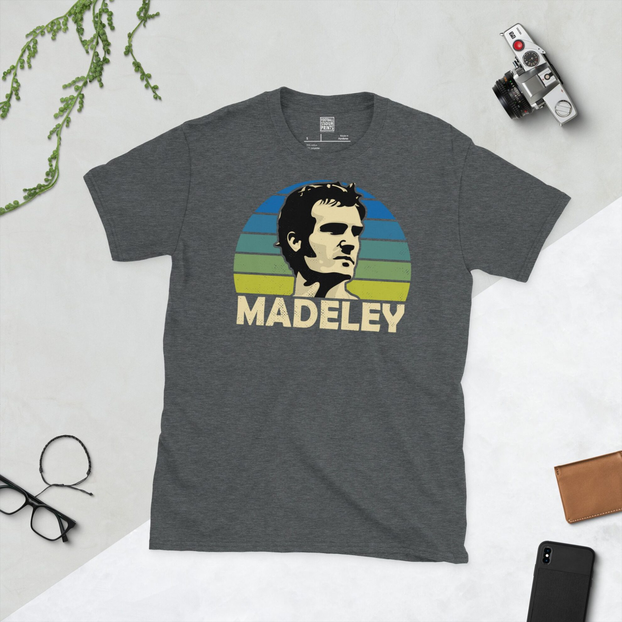 A dark heather T-shirt showing a clean, modern portrait of Paul Madeley, the Leeds icon, with "MADELEY" printed in light yellow below the image on a blue to yellow faded sunburst background.