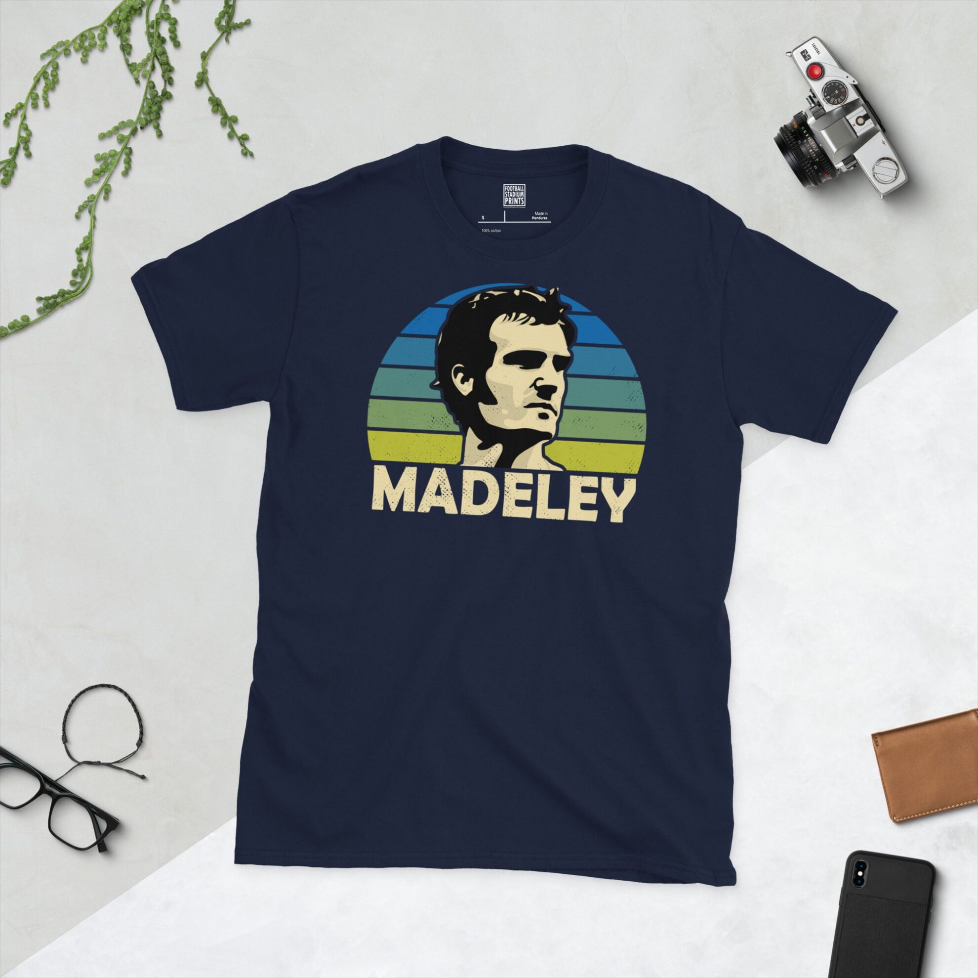 A navy T-shirt displaying an artistic illustration of Paul Madeley, the former Leeds United player. The graphic is backed by a blue to yellow faded sunburst and the name "MADELEY" appears underneath in light yellow.