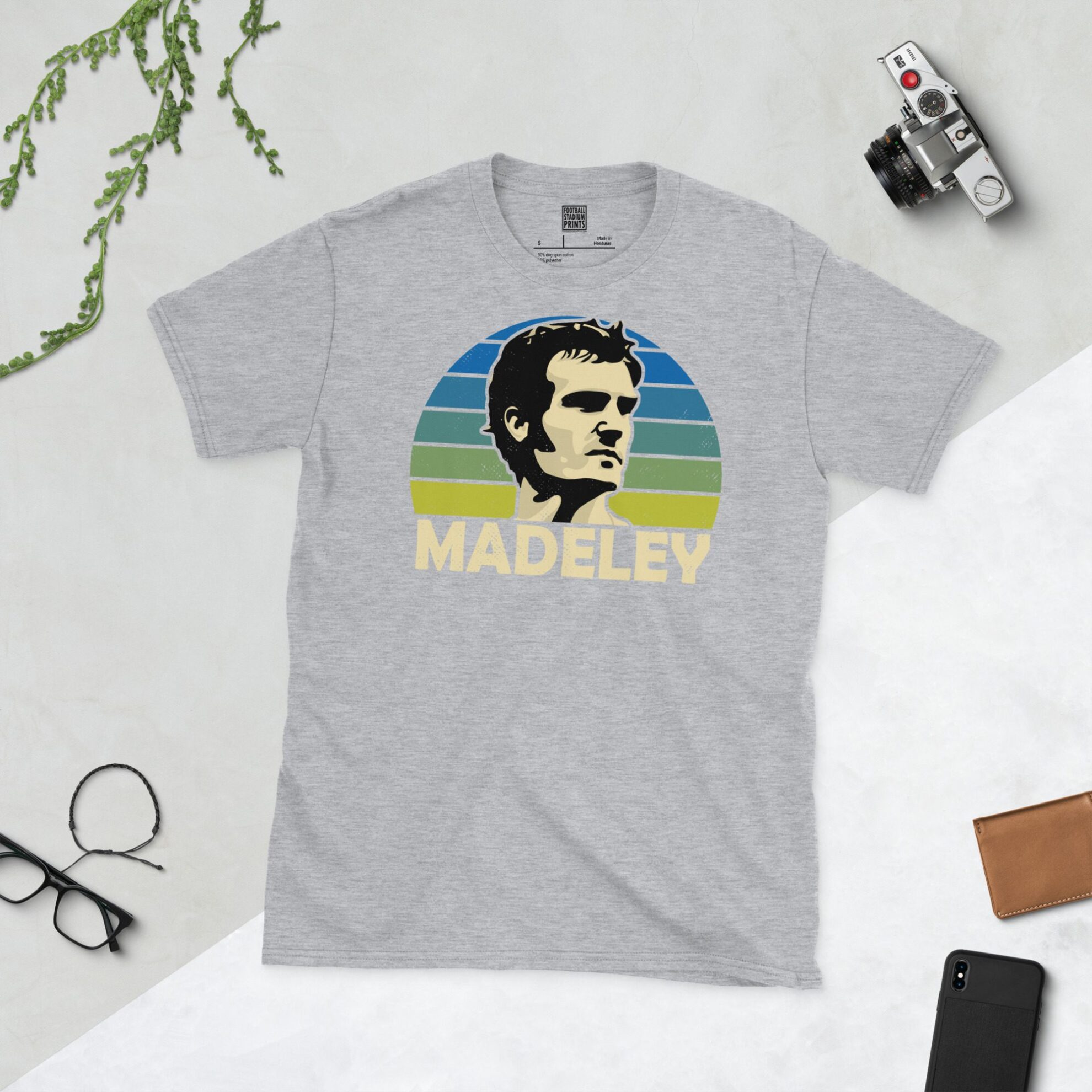 A sports grey T-shirt featuring a unique graphic of Leeds United's Paul Madeley. The image is backed by a blue to yellow faded sunburst and the text "MADELEY" is printed in light yellow below the portrait.
