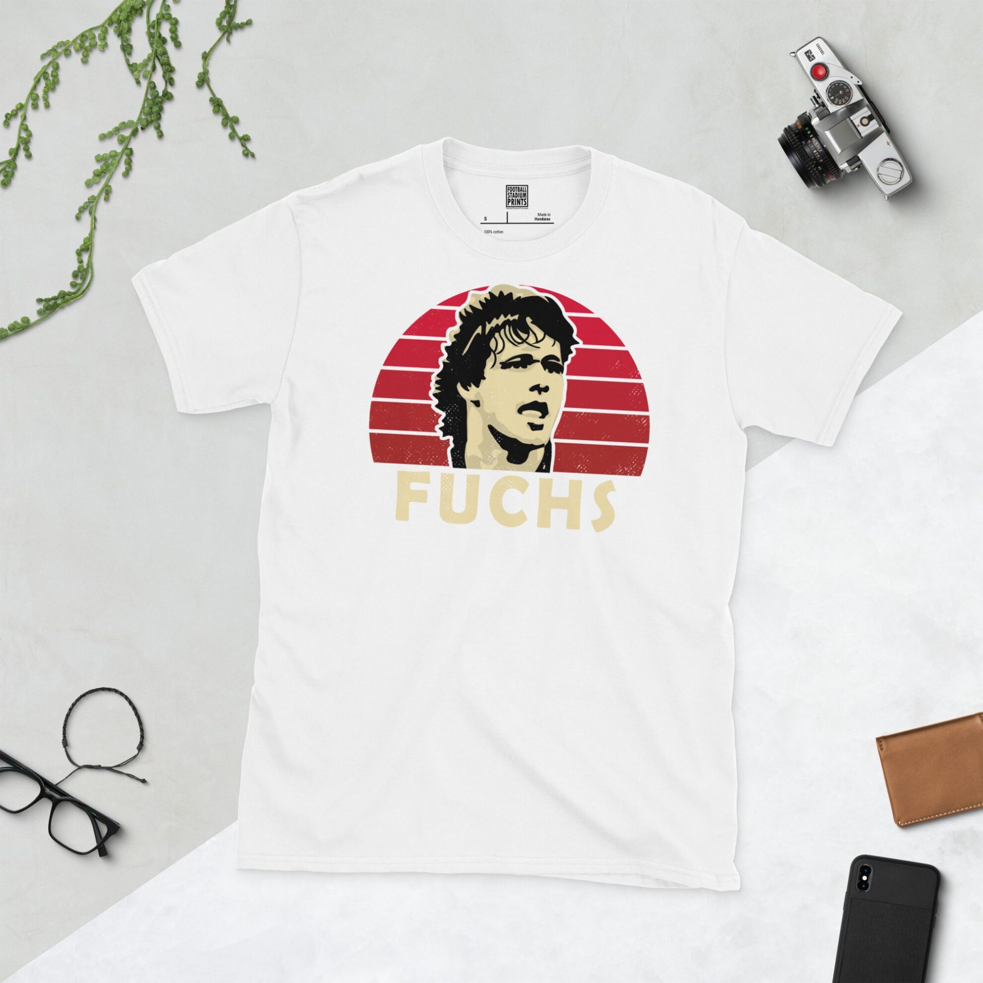A white T-shirt with a bold portrait of football legend Uwe Fuchs. The image is set against a red sunburst background with the name "FUCHS" in light yellow underneath.
