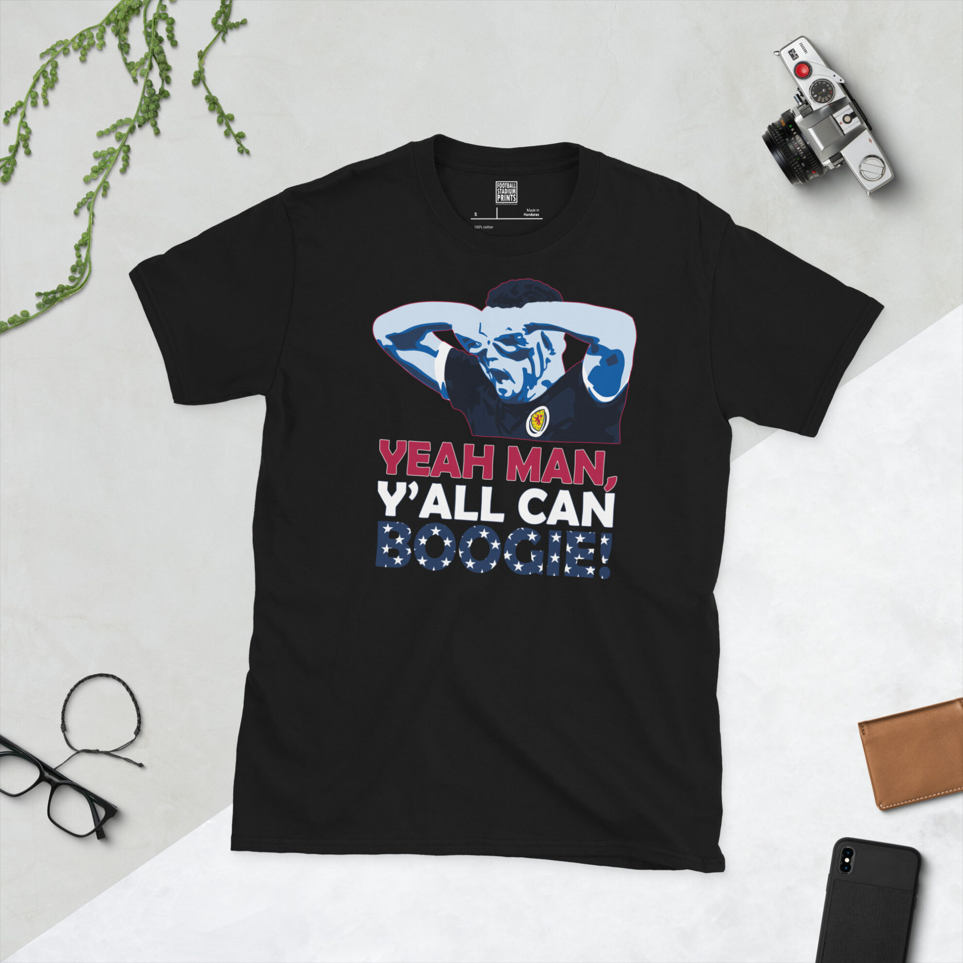 A black T-shirt featuring a graphic of Scotland star John McGinn performing his "specs" celebration. The quote "YEah man, y'all can boogie" is printed on the design, celebrating the World Cup 2026 qualification in North America.