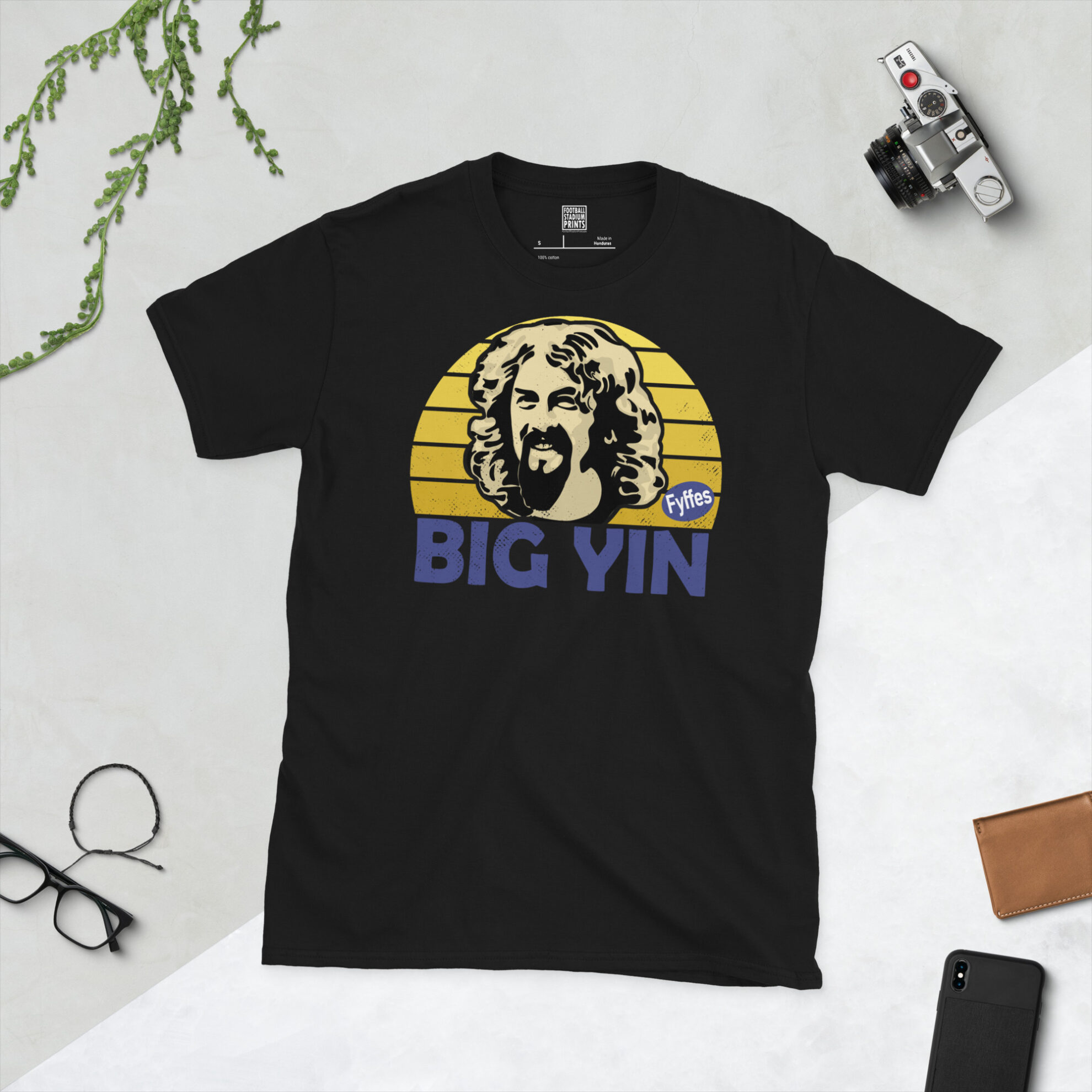 A black T-shirt featuring a stylised, distressed graphic of comedian Billy Connolly. The portrait is backed by a yellow sunburst referencing his banana boots, with the text "BIG YIN" printed in purple below.