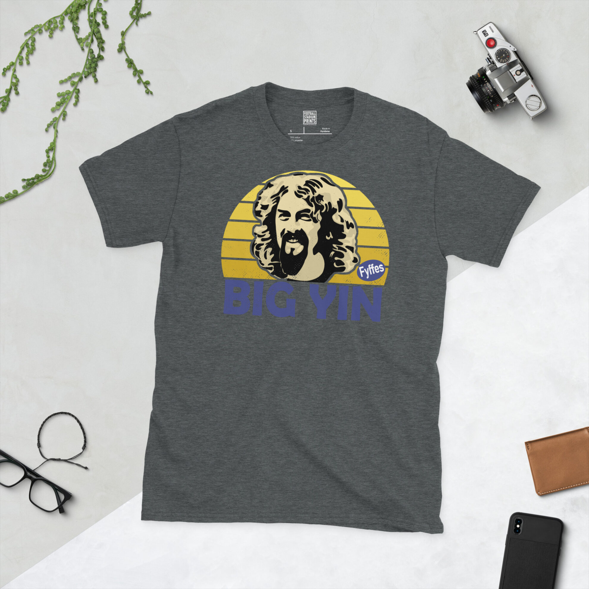 A dark heather T-shirt showing a clean, modern portrait of Billy Connolly, the comedy icon, with "BIG YIN" printed in purple below the image on a yellow sunburst background.
