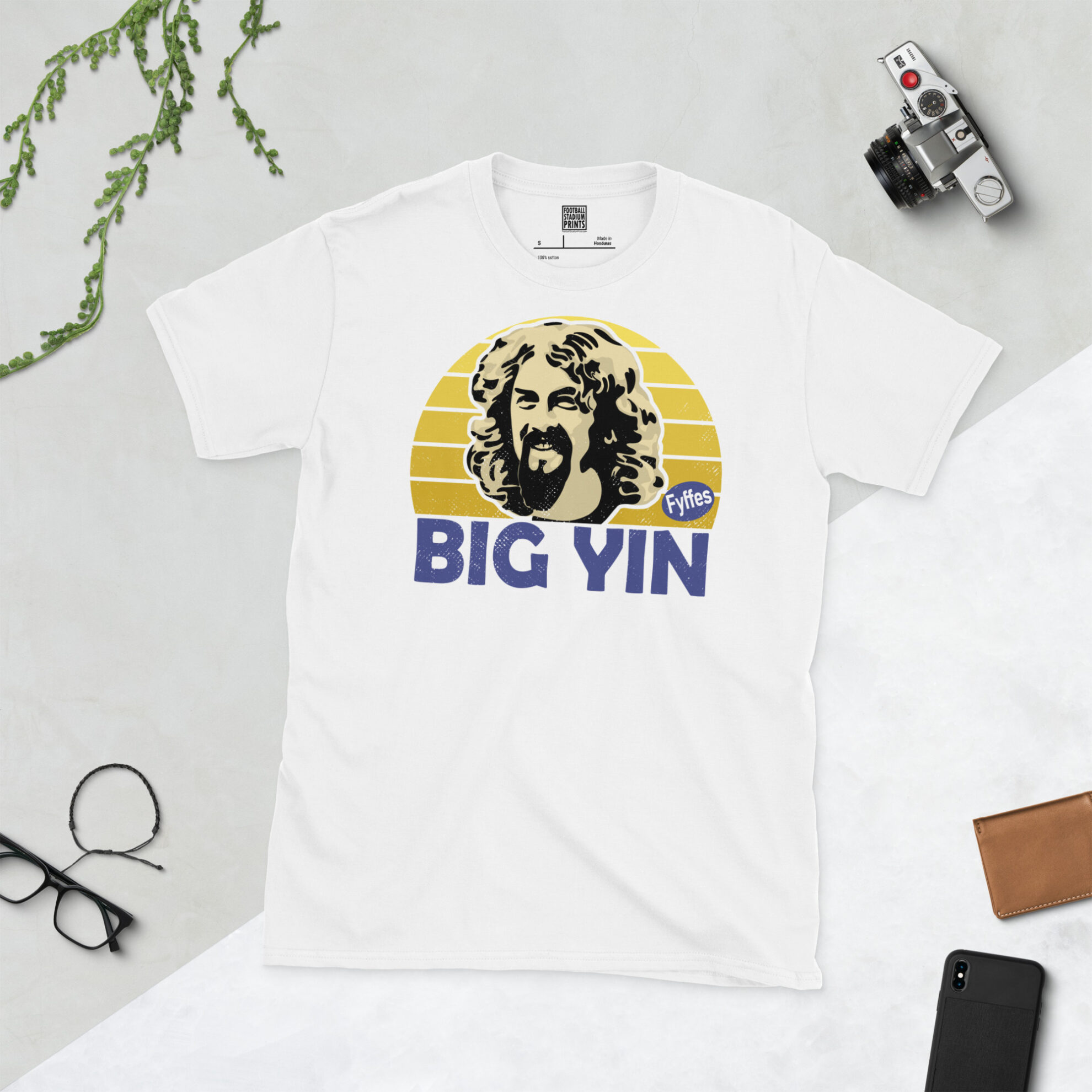 A white T-shirt with a bold, retro graphic of Scottish comedy legend Billy Connolly. The image is set against a yellow sunburst background and the name "BIG YIN" is in purple underneath.