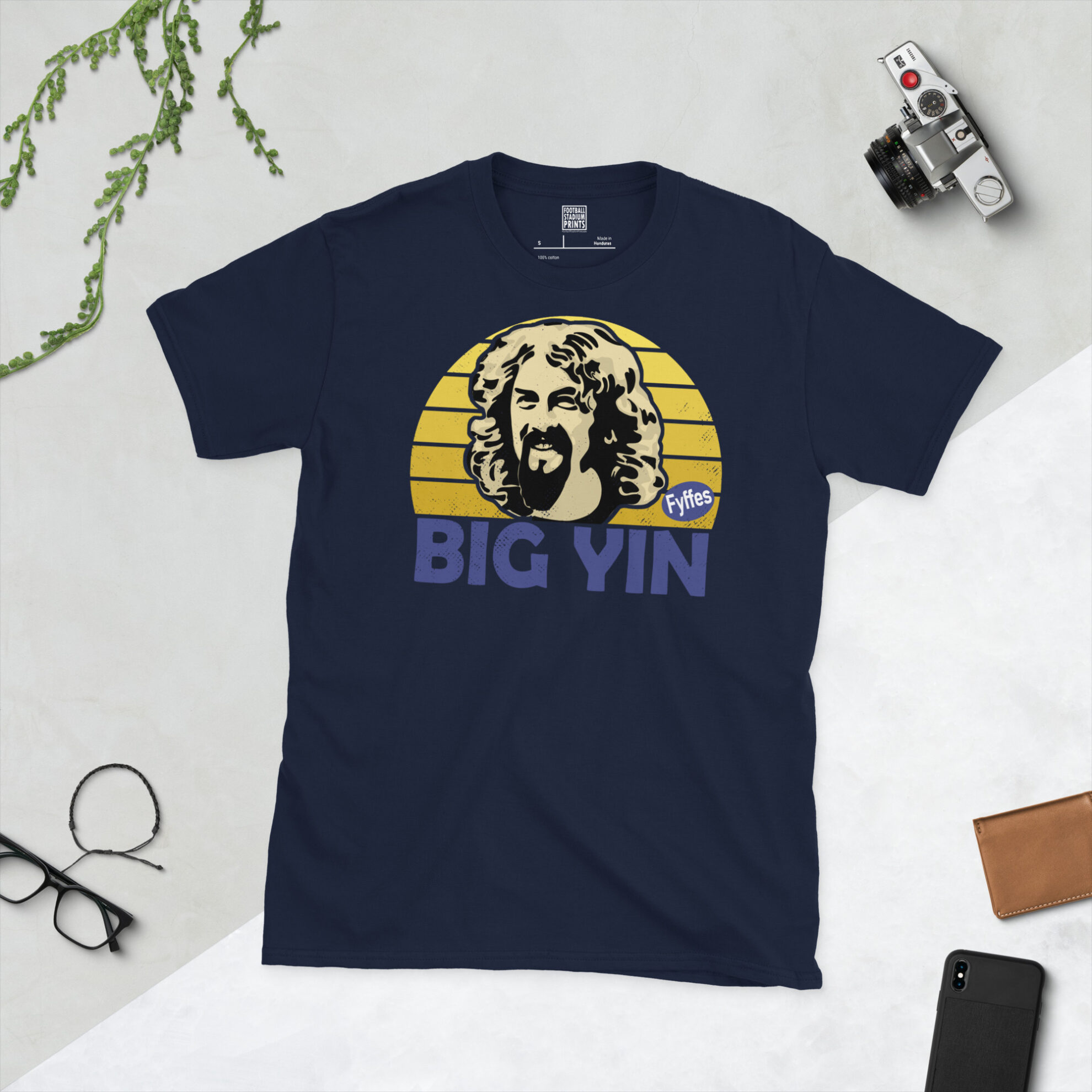 A navy T-shirt displaying an artistic illustration of Billy Connolly, "The Big Yin". The graphic features a yellow striped sunburst and the nickname "BIG YIN" appears underneath in purple.