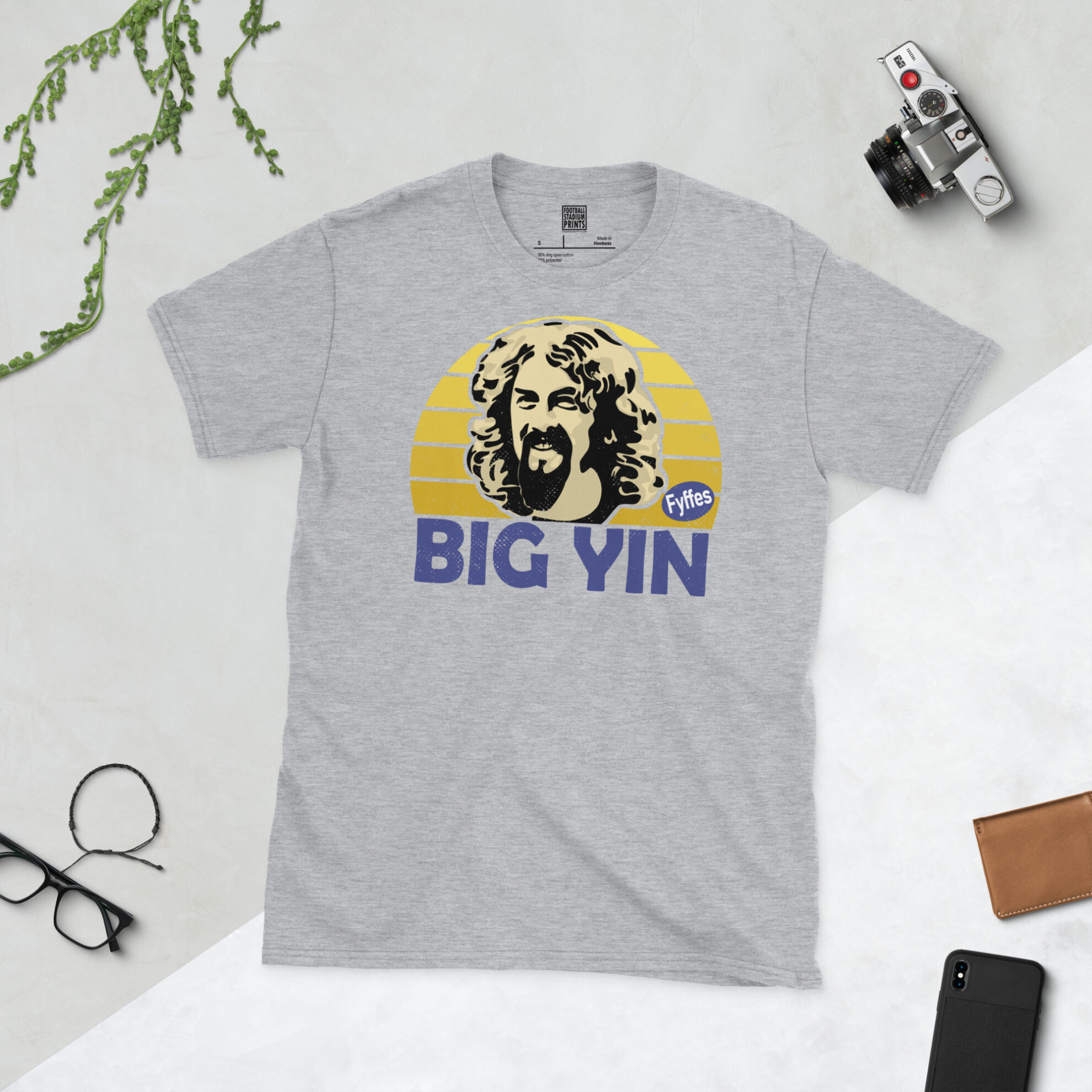 A sports grey T-shirt featuring a unique graphic of Billy Connolly. The image is backed by a yellow sunburst and the text "BIG YIN" is printed below the portrait in purple.