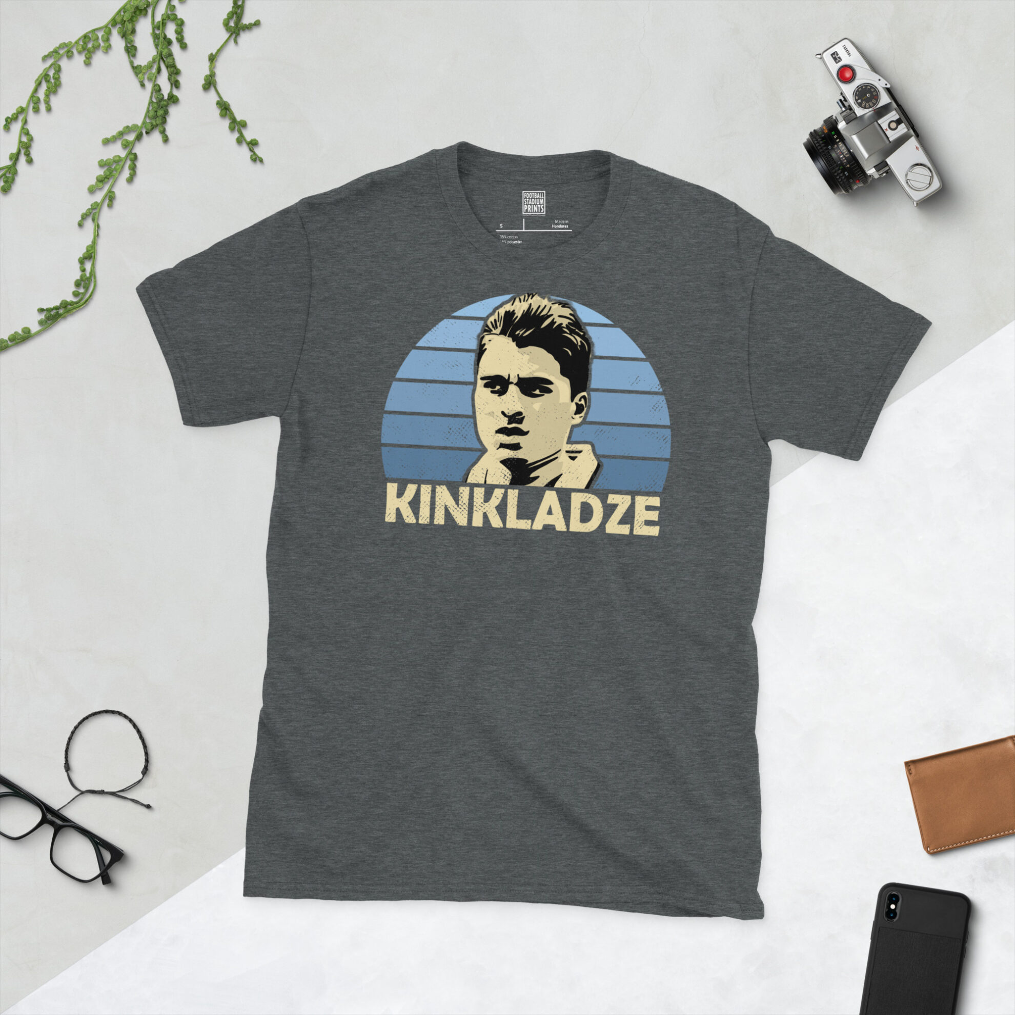 A dark heather T-shirt showing a clean, modern portrait of Georgi Kinkladze, the City icon, with "KINKLADZE" printed in light yellow below the image on a blue sunburst background.