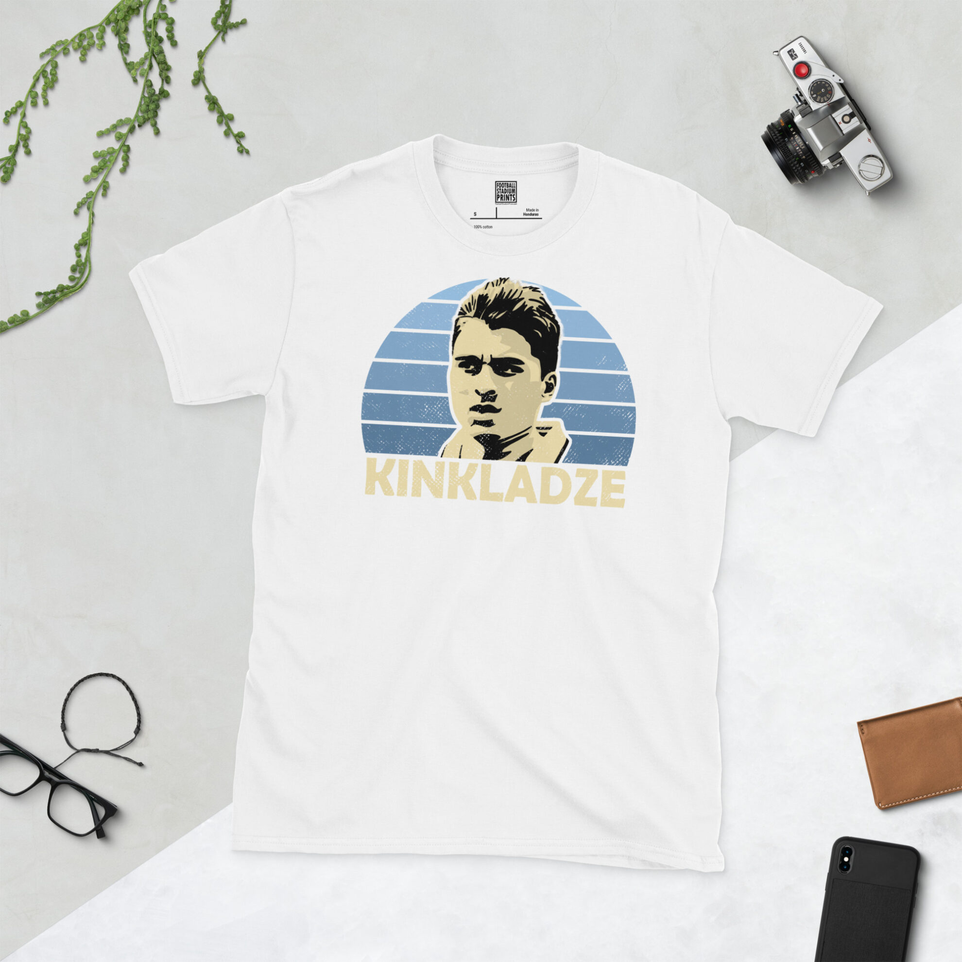 A white T-shirt with a bold, retro graphic of football legend Georgi Kinkladze. The image is set against a blue sunburst background and the name "KINKLADZE" is in light yellow underneath.
