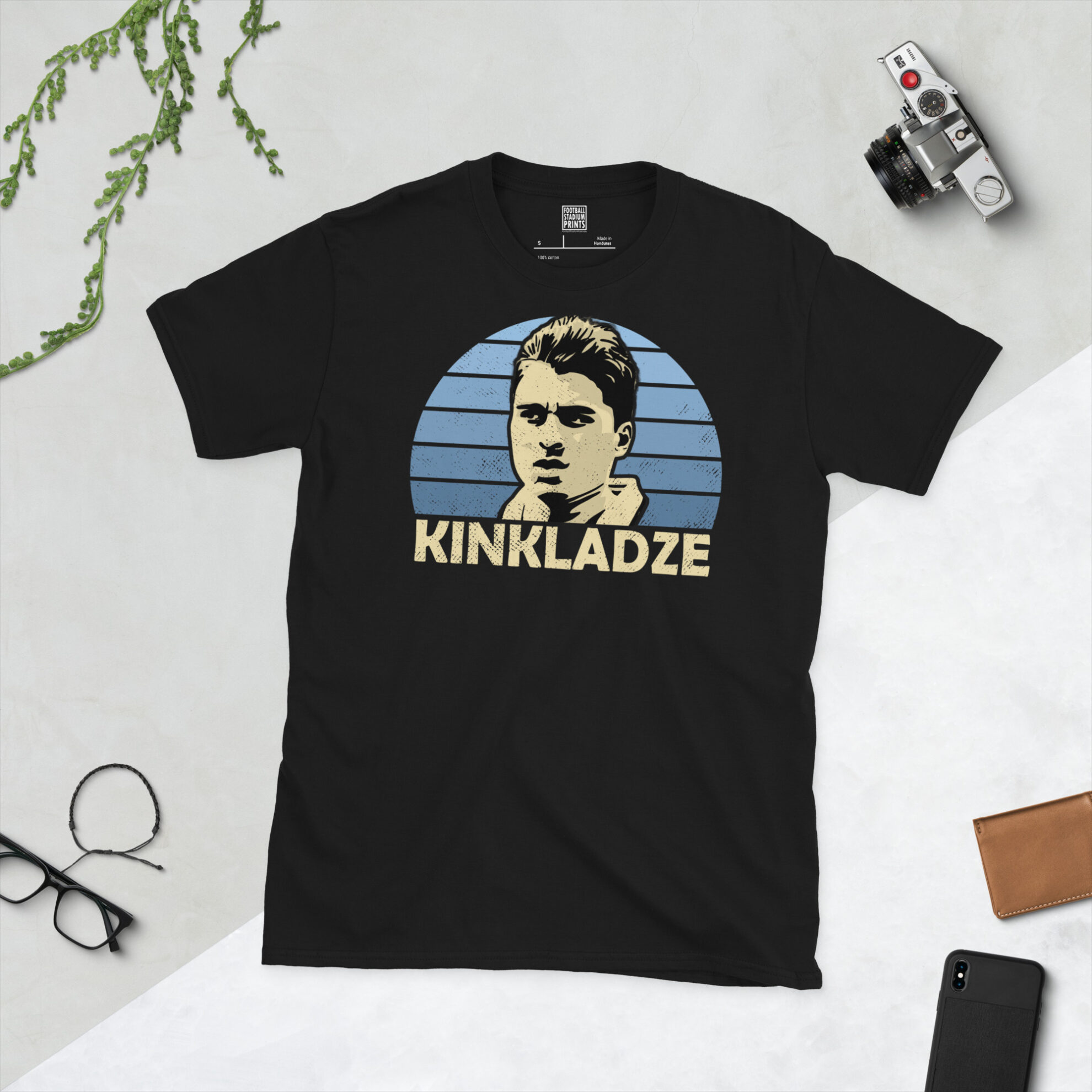 A black T-shirt featuring a stylised, distressed graphic of Manchester City legend Georgi Kinkladze. The portrait is backed by a blue striped sunburst, with the name "KINKLADZE" printed in light yellow below.