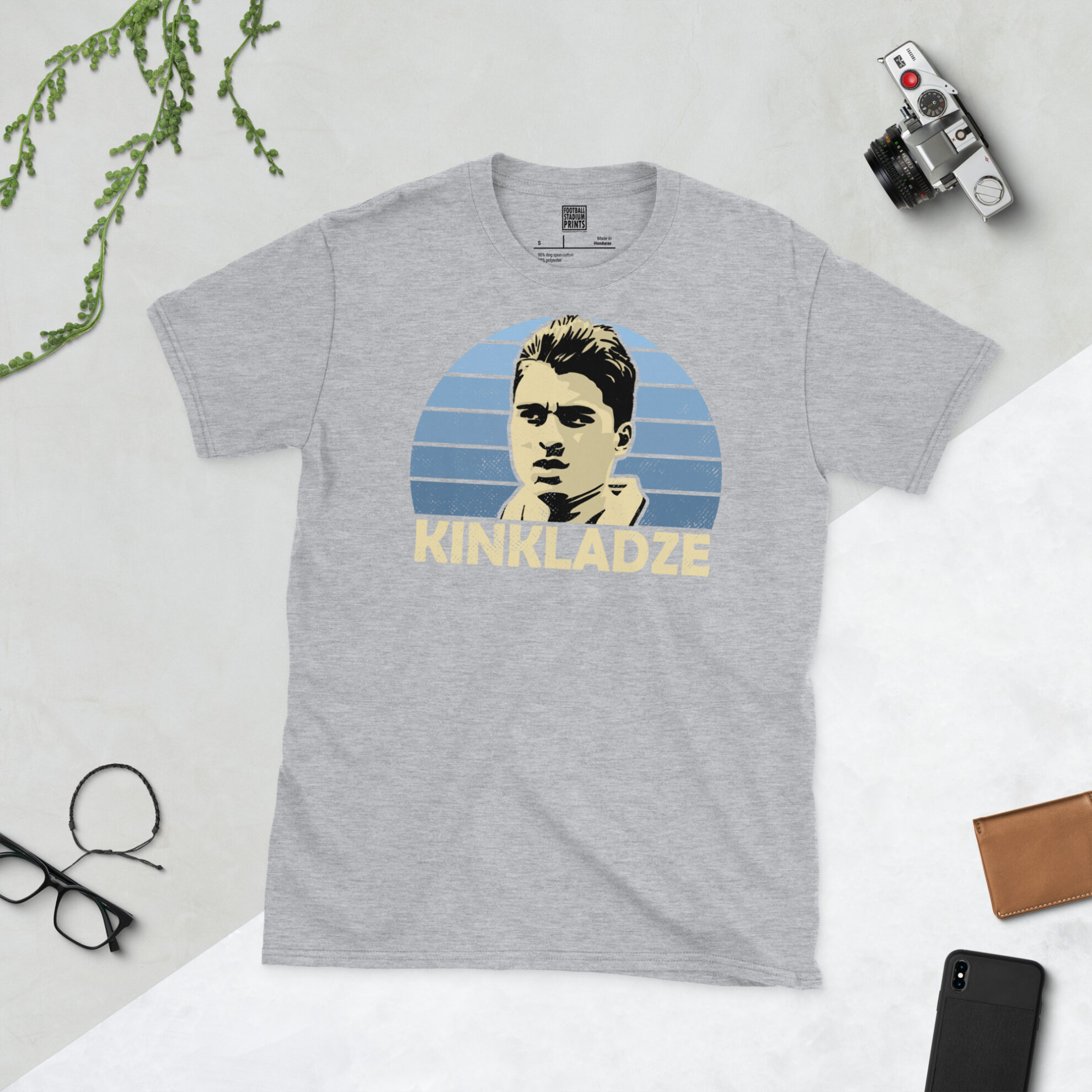 A sports grey T-shirt featuring a unique graphic of Georgi Kinkladze. The image is backed by a blue sunburst and the text "KINKLADZE" is printed below the portrait in light yellow.