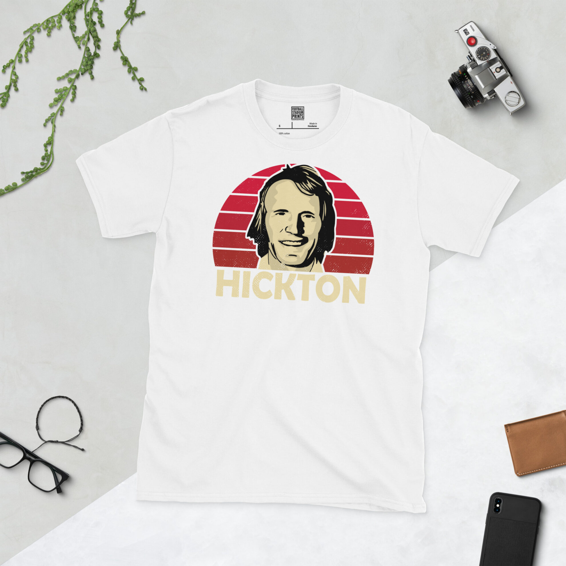 A white T-shirt with a bold, retro graphic of Middlesbrough star John Hickton. The image is set against a red sunburst background and the name "HICKTON" is in light yellow underneath.