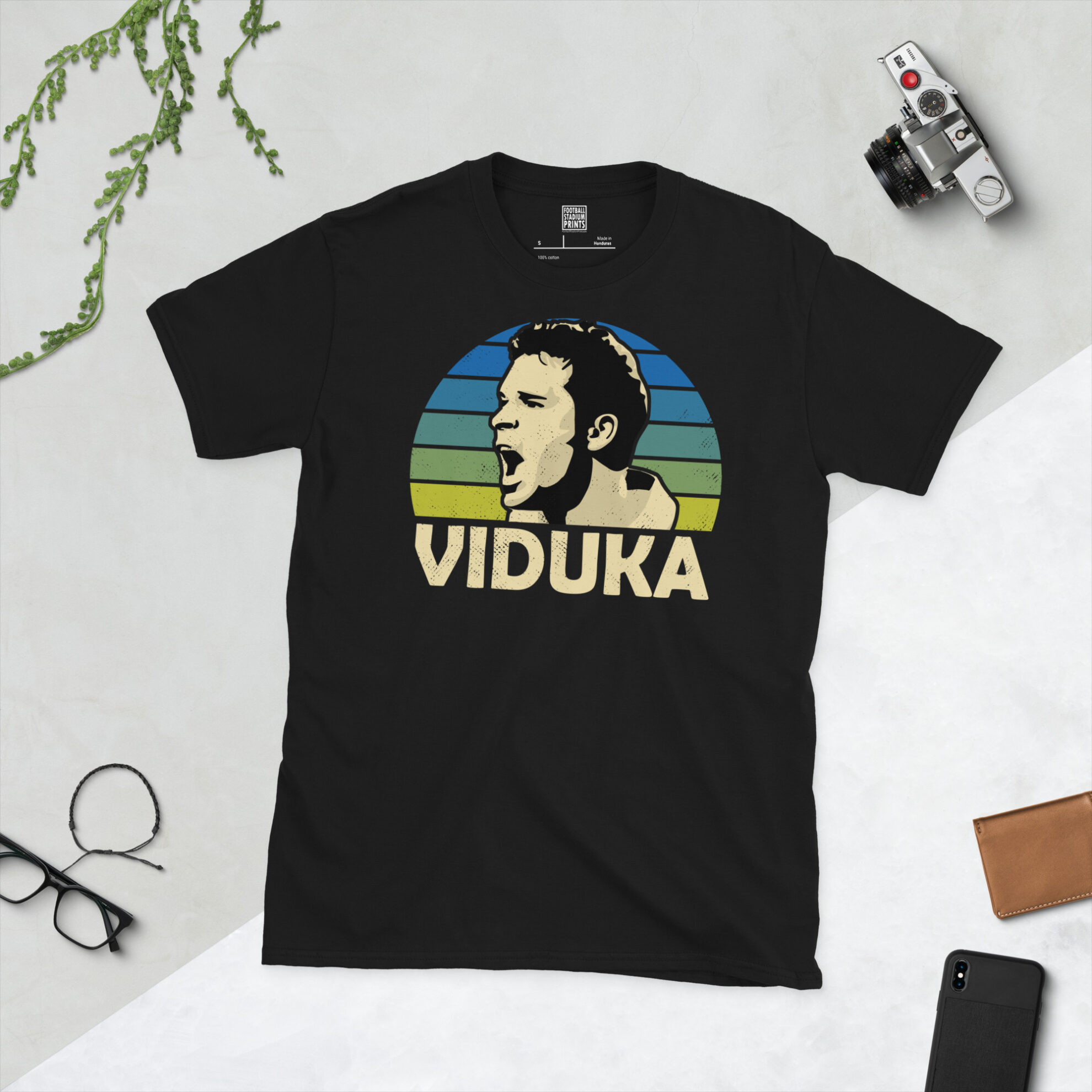 A black T-shirt featuring a stylised, distressed graphic of Leeds United icon Mark Viduka. The portrait is backed by a blue to yellow fade sunburst, with the name "VIDUKA" printed in white below.