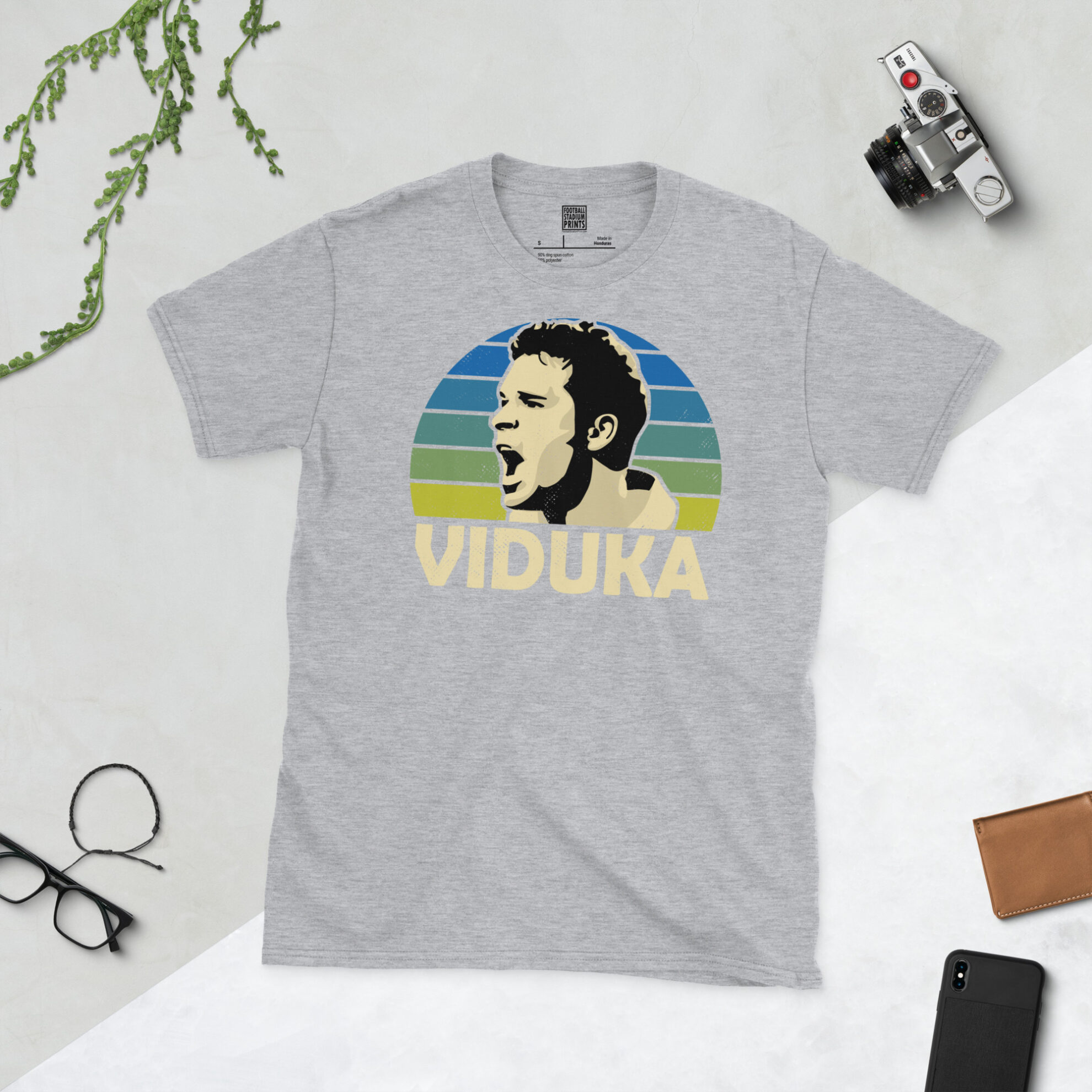 A sports grey T-shirt featuring a unique graphic of Mark Viduka. The image is backed by a blue to yellow sunburst and the text "VIDUKA" is printed below the portrait in white.