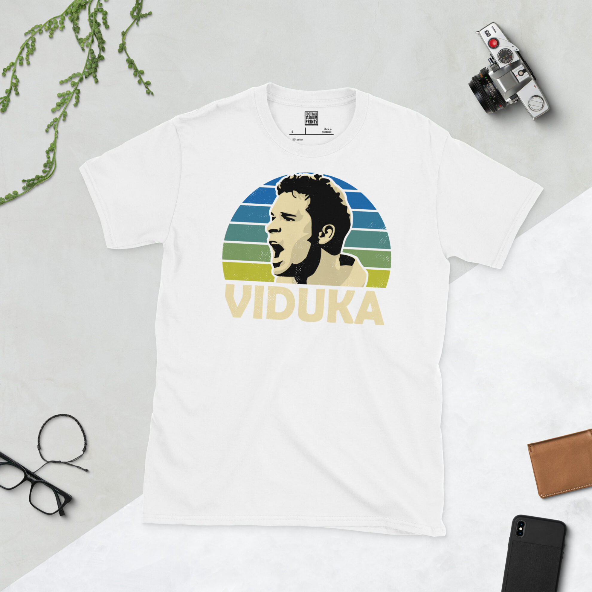 A white T-shirt with a bold, retro graphic of football legend Mark Viduka. The image is set against a blue to yellow fade sunburst background and the name "VIDUKA" is in white underneath.
