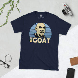 A navy T-shirt displaying an artistic illustration of Shaun Goater, the legendary Man City striker. The graphic features a blue sunburst and the title "THE GOAT" appears underneath in light yellow.