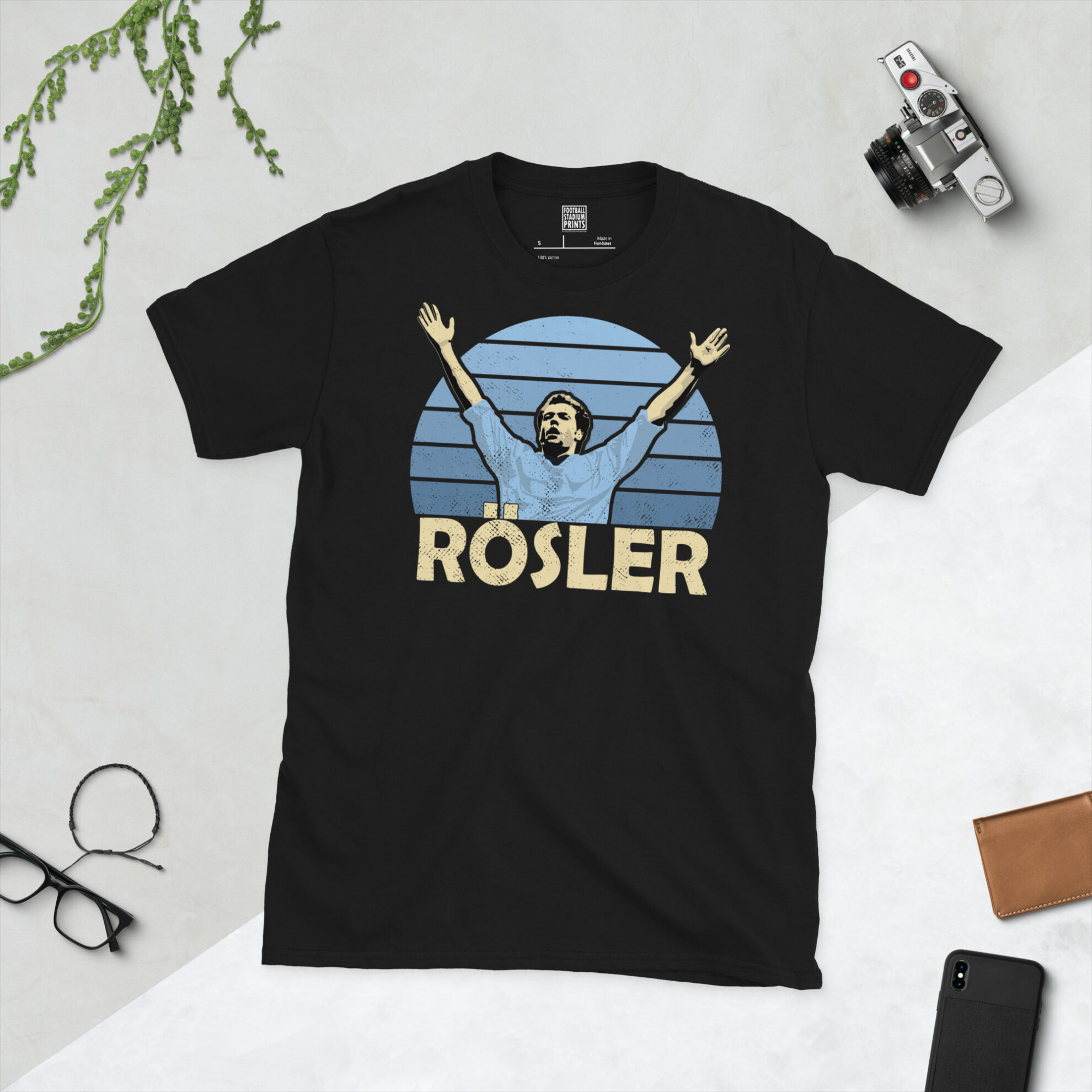 A black T-shirt featuring a stylised, distressed graphic of Manchester City legend Uwe Rosler. The portrait is backed by a blue striped sunburst, with the name "ROSLER" printed in light yellow below.