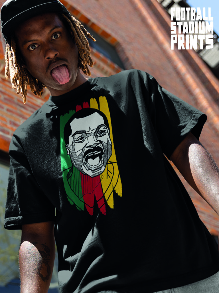 A man wearing a black t-shirt featuring a vibrant graphic of Cameroon legend Roger Milla, highlighted by the green, red, and yellow colors of the national flag.