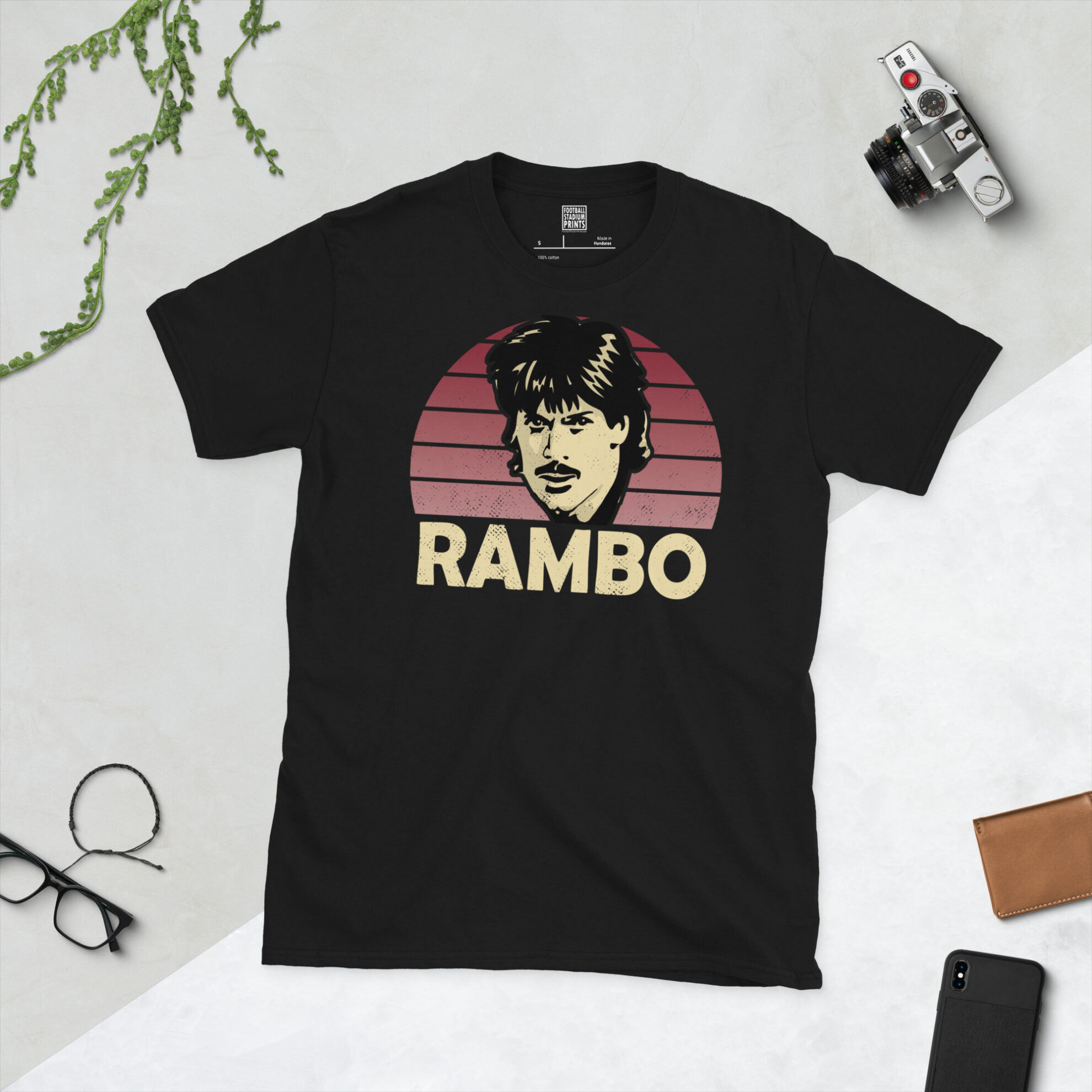 A premium black T-shirt featuring a distressed claret sunburst graphic of Northampton Town legend Graham Reed, with "RAMBO" typography below.