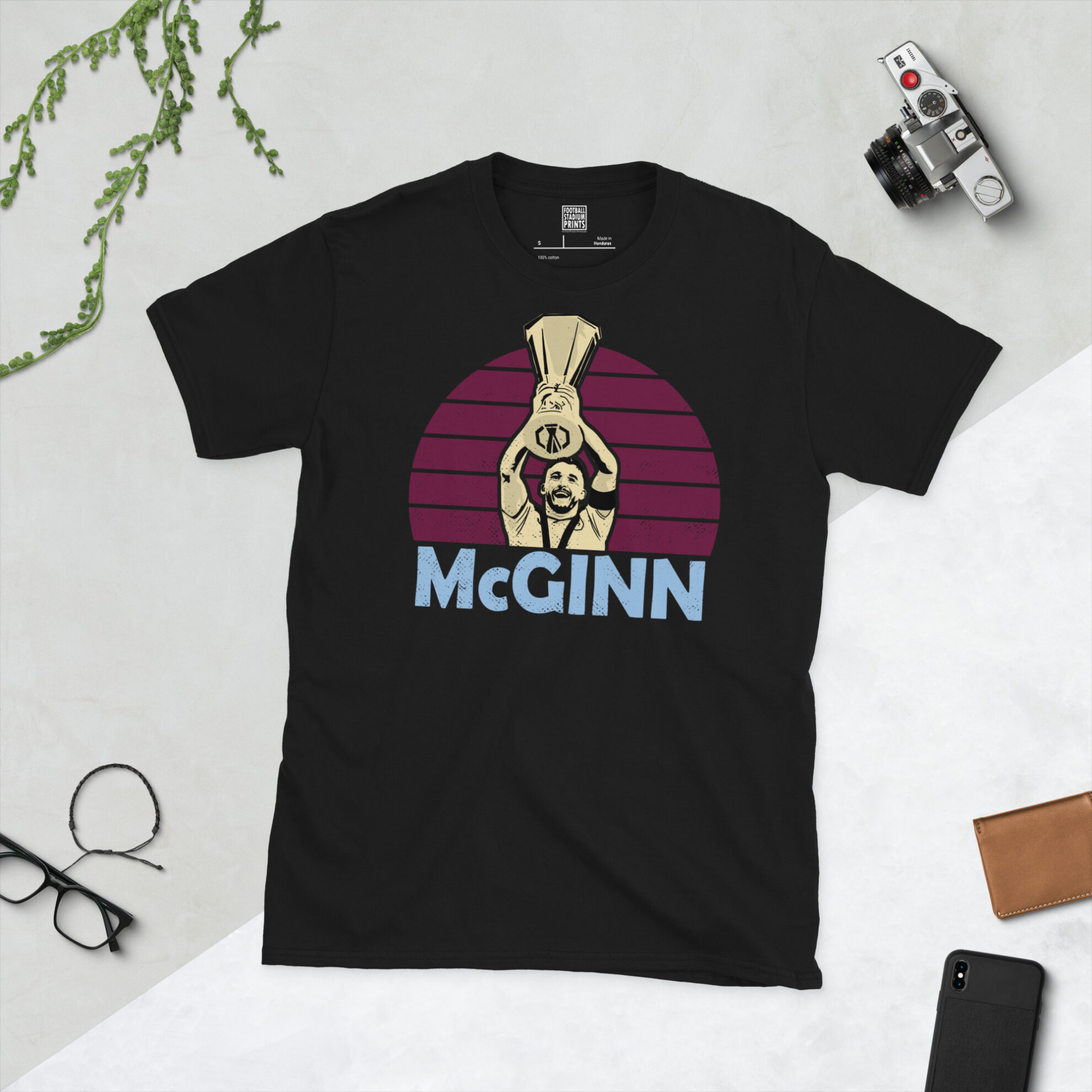 A premium black T-shirt featuring a distressed claret sunburst graphic of Aston Villa captain John McGinn lifting the 2026 Europa League trophy with "McGINN" in sky blue.