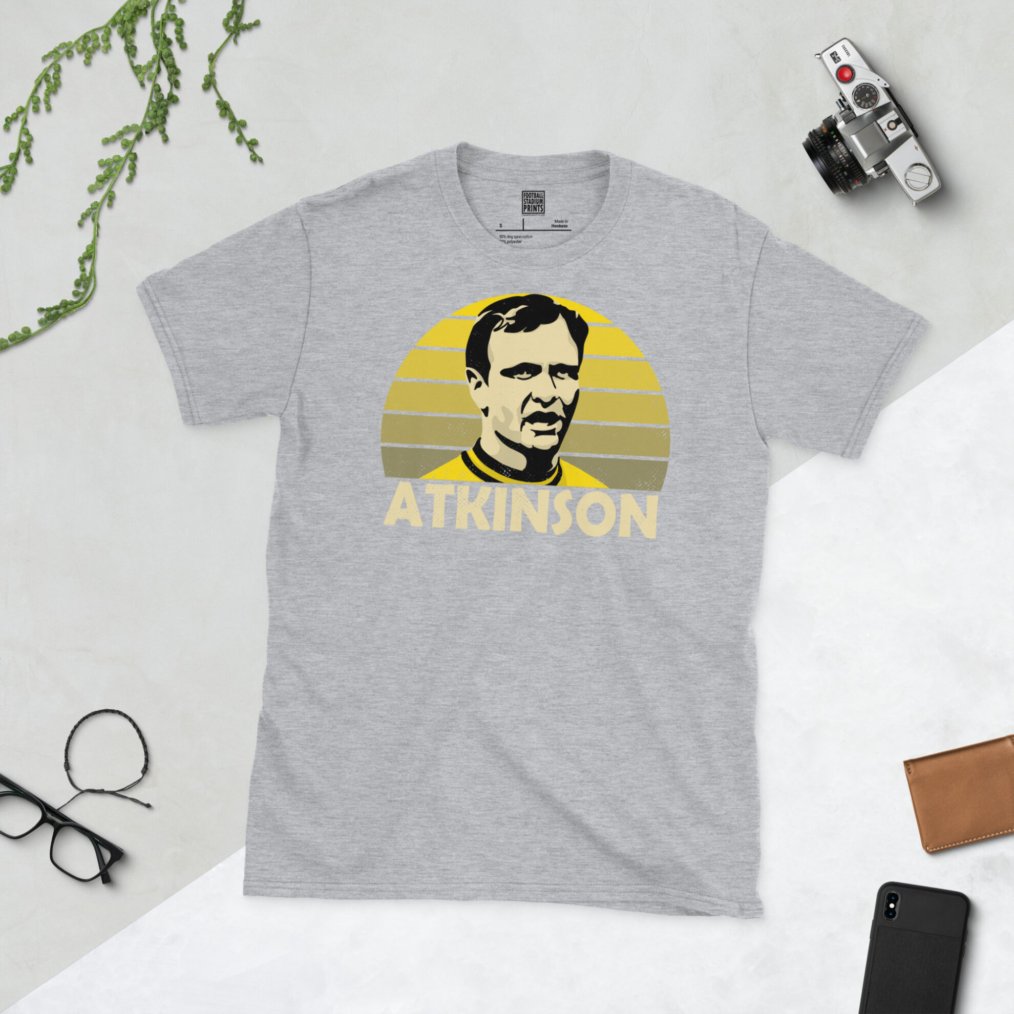 A sports grey T-shirt featuring a bold yellow-to-blue sunburst design of Ron Atkinson, finished with his name in a classic retro font.