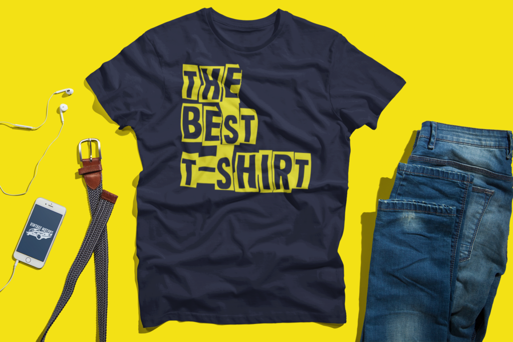 Flat lay of navy blue t-shirt printed with bold yellow stencil block typography reading The Best T-Shirt on a vibrant yellow background with jeans and accessories